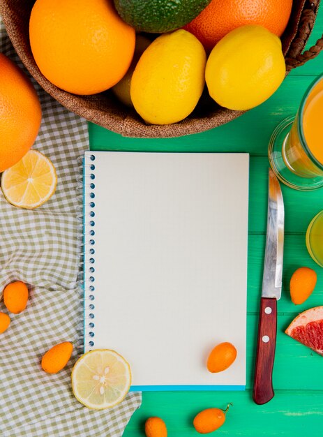 Vista superiore del blocco note con il coltello e il succo arancio del kumquat del limone intorno su fondo verde con lo spazio della copia