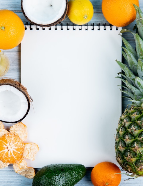 Vista superiore del blocco note con il limone arancio dell'ananas dell'avocado del mandarino della noce di cocco intorno su fondo di legno con lo spazio della copia