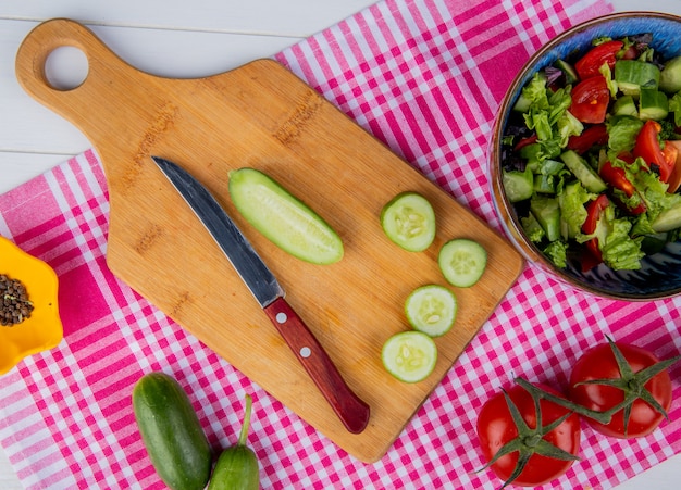 Vista superiore del cetriolo tagliato e affettato con il coltello sul tagliere e pepe nero del pomodoro dell'insalata di verdure sul panno del plaid e sulla superficie di legno