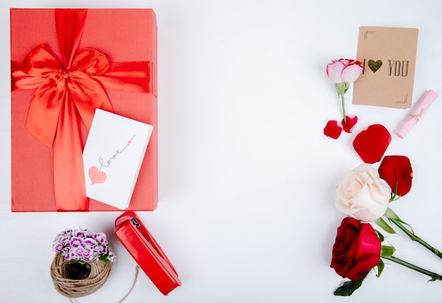 Vista superiore del contenitore di regalo rosso con un arco e rose rosse e bianche di colore con la cucitrice meccanica e la piccola cartolina su fondo bianco con lo spazio della copia