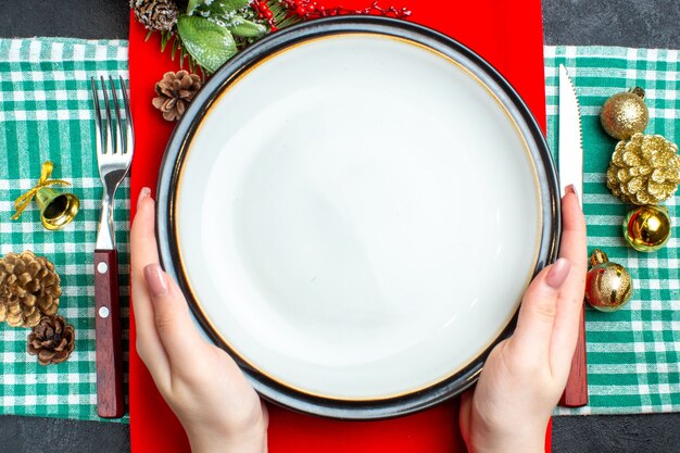 Vista superiore del fondo nazionale del pasto di Christmal con la mano che tiene gli accessori della decorazione delle posate dei piatti vuoti messi sull'asciugamano spogliato verde