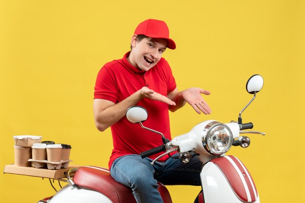 Vista superiore del giovane adulto sorridente che indossa la camicetta rossa e il cappello che consegna gli ordini che indicano lo scooter sulla parete gialla