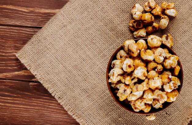 Vista superiore del popcorn al caramello dolce in una ciotola di legno su tela di sacco su fondo rustico con spazio di copia