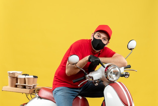 Vista superiore del ragazzo curioso del corriere che indossa guanti di camicetta e cappello rossi nella mascherina medica che consegna l'ordine che si siede sul motorino