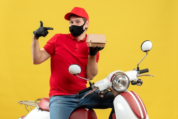 Vista superiore del ragazzo di consegna fiducioso che indossa guanti di camicetta e cappello rossi in maschera medica che si siede sullo scooter che mostra l'ordine