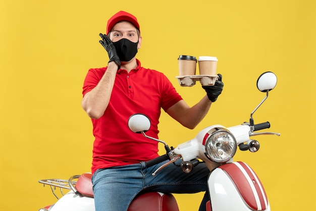 Vista superiore del ragazzo di consegna fiducioso sorridente che indossa guanti uniformi e cappello in maschera medica che si siede sullo scooter che mostra gli ordini