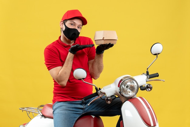 Vista superiore del ragazzo di consegna soddisfatto fiducioso che indossa guanti di camicetta e cappello rossi nella mascherina medica che si siede sullo scooter che mostra l'ordine