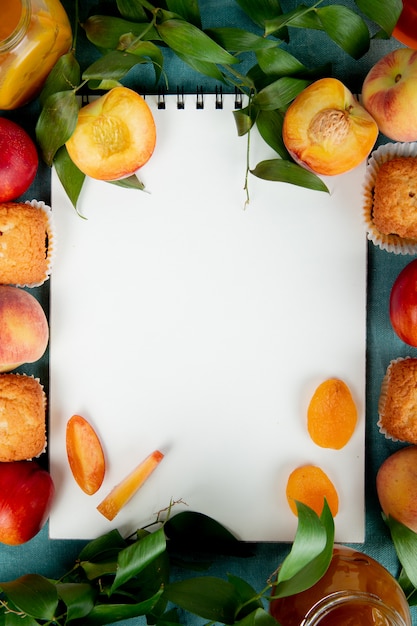 Vista superiore dell'inceppamento della pesca dei cupcakes delle pesche con il blocco note sul centro su superficie blu decorata con le foglie con lo spazio della copia