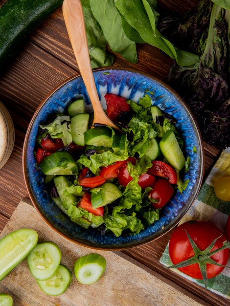 Vista superiore dell'insalata di verdure con il pomodoro tagliato e affettato del basilico degli spinaci del cetriolo su superficie di legno