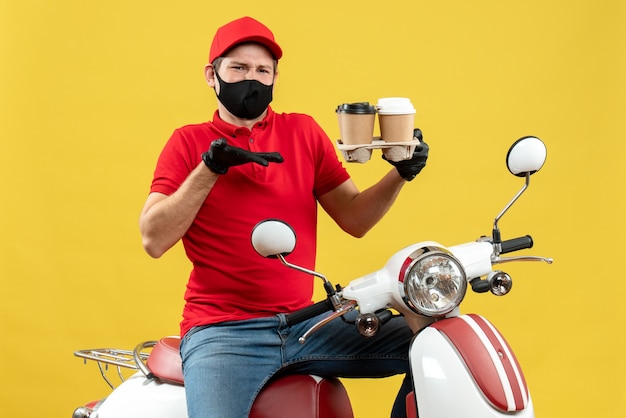 Vista superiore dell'uomo di consegna confuso che indossa guanti uniformi e cappello in maschera medica che si siede sullo scooter che mostra gli ordini