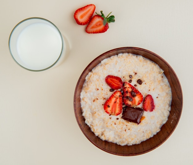 Vista superiore della ciotola di farina d'avena con cioccolato e fragole della ricotta con bicchiere di latte su superficie bianca