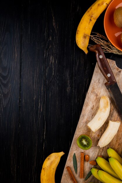 Vista superiore della frutta della banana con la mandorla, i bastoncini di cannella e il vecchio coltello da cucina su un tagliere di legno sul nero con lo spazio della copia