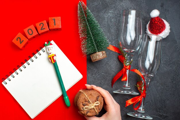 Vista superiore della mano che tiene il taccuino a spirale e i calici di vetro dell'albero di Natale numeri mano che tiene i biscotti impilati su fondo scuro e rosso