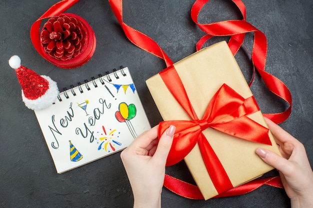 Vista superiore della mano che tiene un cono di conifera regalo con nastro rosso e taccuino con la scrittura del nuovo anno e il cappello di Babbo Natale su sfondo scuro