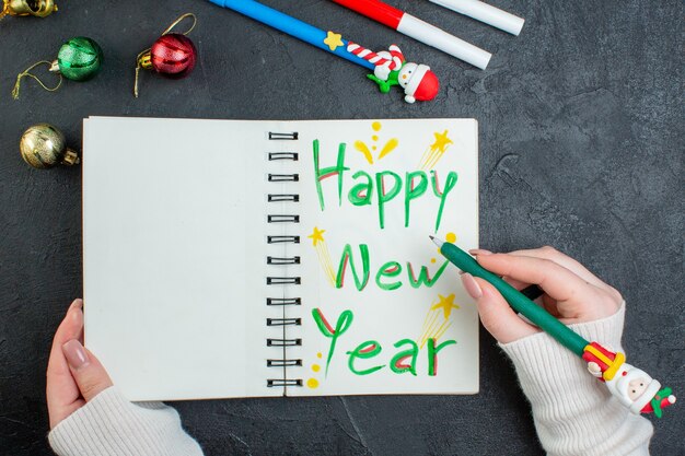 Vista superiore della mano che tiene una penna sul taccuino a spirale con accessori di decorazione di scrittura di felice anno nuovo sulla tavola nera