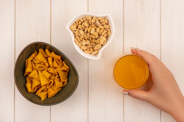 Vista superiore della mano femminile che tiene un bicchiere di succo d'arancia con una ciotola di snack di mais a forma di cono con pinoli su una ciotola bianca su un tavolo di legno beige