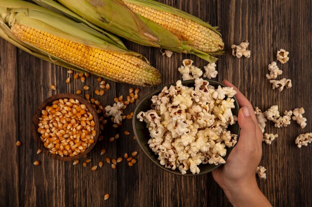 Vista superiore della mano femminile che tiene una ciotola di popcorn con chicchi di mais su una ciotola di legno con mais fresco su un tavolo di legno