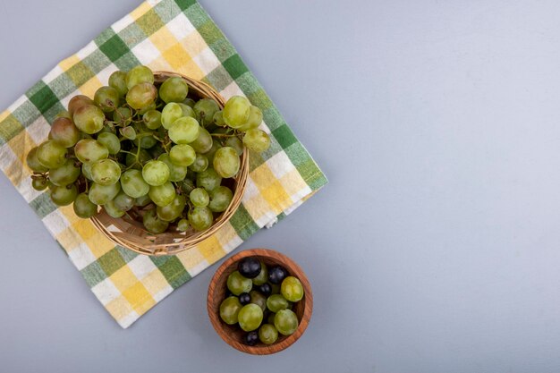 Vista superiore della merce nel cestino dell'uva bianca sul panno del plaid e degli acini dell'uva nella ciotola su fondo grigio con lo spazio della copia