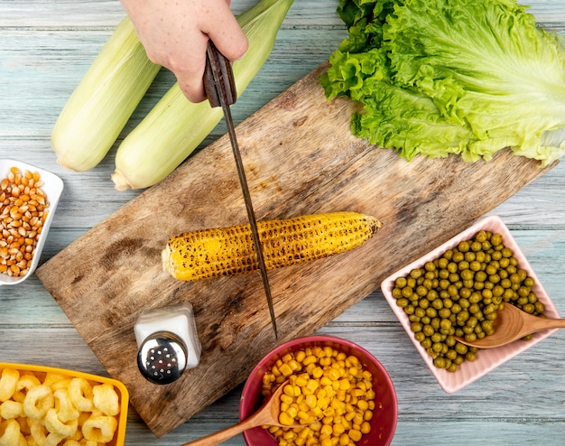 Vista superiore della pannocchia di mais del taglio manuale della donna con il coltello sul tagliere e semi del cereale del sale della lattuga dei piselli su superficie di legno