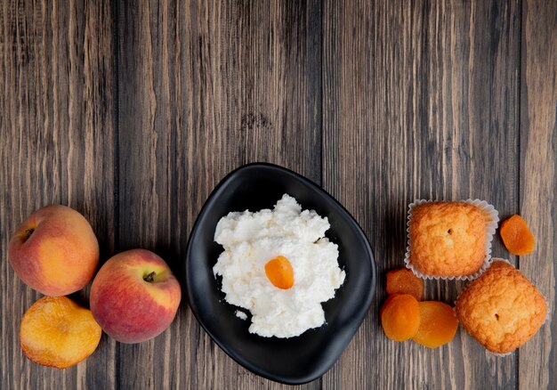 Vista superiore della ricotta in una ciotola nera con le pesche fresche dei muffin e le albicocche secche su legno rustico con lo spazio della copia