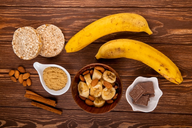 Vista superiore delle banane affettate con la mandorla in una ciotola di legno e le banane mature fresche con i cracker del riso e del cioccolato su legno