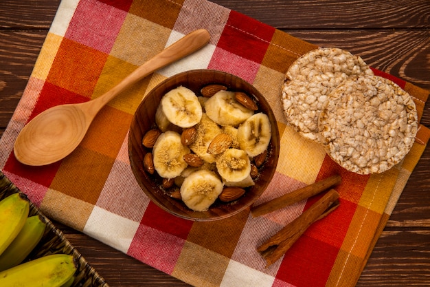 Vista superiore delle banane affettate con la mandorla in una ciotola e cucchiai di legno con i cracker del riso e bastoncini di cannella su rustico