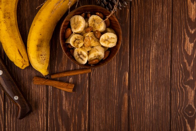 Vista superiore delle banane affettate con la mandorla in una ciotola e delle banane mature fresche con i bastoni di cannella su legno con lo spazio della copia