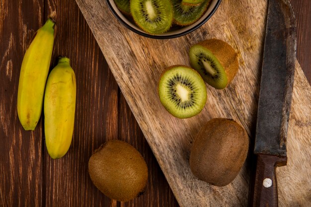 Vista superiore delle fette di kiwi in una ciotola sul bordo di legno e delle banane fresche con un coltello su rustico