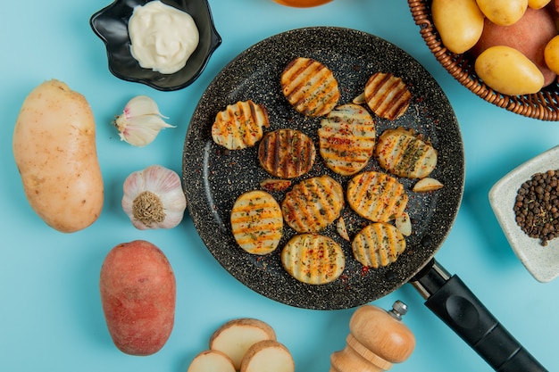 Vista superiore delle fette fritte della patata in padella con pepe nero del sale dell'aglio della maionese della merce nel carrello cruda sul blu