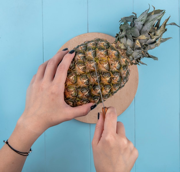 Vista superiore delle mani della donna che tagliano ananas sul tagliere e sulla superficie del blu