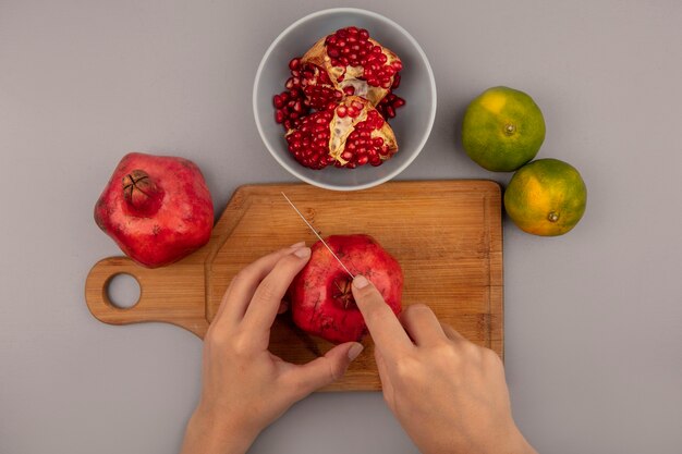 Vista superiore delle mani femminili che tagliano i melograni rossi freschi su una tavola di cucina in legno con il coltello con il melograno aperto su una ciotola con i mandarini isolati