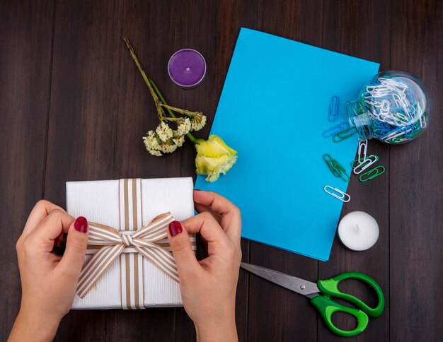 Vista superiore delle mani femminili che tengono il contenitore di regalo con il nastro dell'arco con rosa gialla su legno con lo spazio della copia