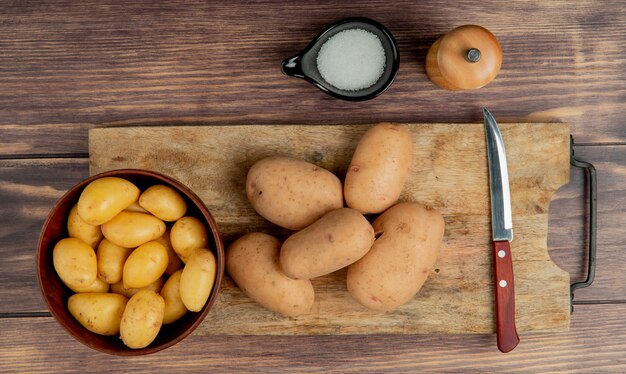 Vista superiore delle patate in ciotola e sul tagliere con coltello e sale su superficie di legno