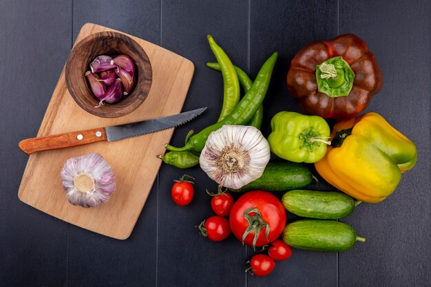 Vista superiore delle verdure come bulbo di aglio e chiodi di garofano con il coltello sul tagliere e cetriolo del pepe del pomodoro su superficie nera