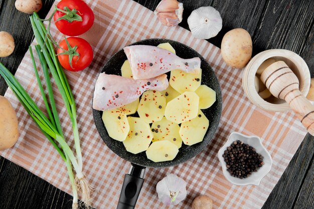 Vista superiore delle verdure come patata dell'aglio dello scalogno del pomodoro con la pentola di pezzi della patata e della coscia di pollo e pepe nero su fondo di legno