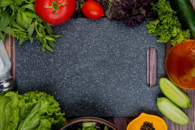 Vista superiore delle verdure intere e tagliate come coriandolo della lattuga del cetriolo della menta del basilico del pomodoro con pepe nero del sale e tagliere su superficie di legno