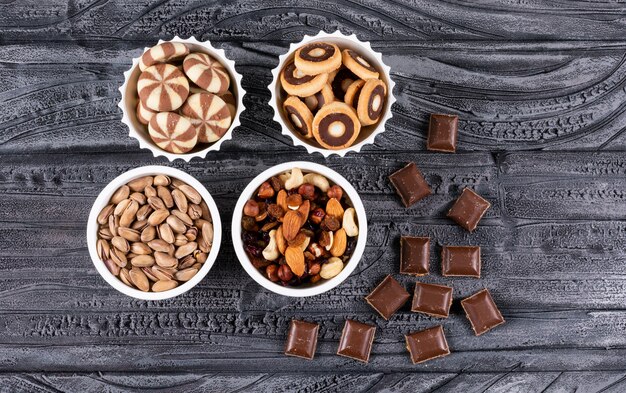 Vista superiore di diversi tipi di snack come noci, biscotti e cioccolato in ciotole su superficie scura orizzontale