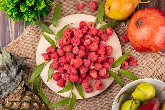 Vista superiore di lamponi con foglie sul tagliere e ananas e melograni su tela di sacco e superficie di legno