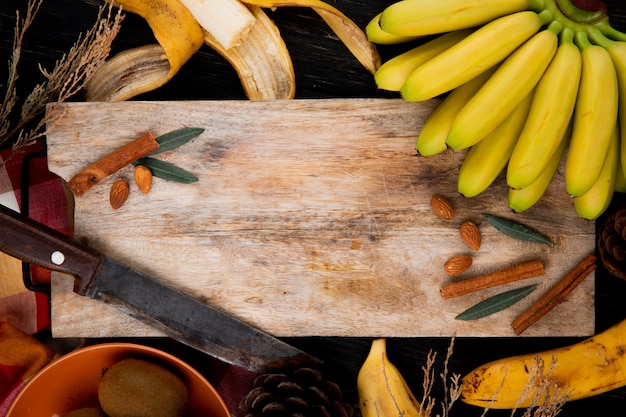 Vista superiore di un mazzo di banana con mandorle, bastoncini di cannella e vecchio coltello da cucina su un tagliere di legno sul nero