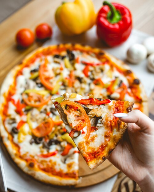 Vista superiore di una mano femminile che tiene un pezzo di pizza con i pomodori e il formaggio dei peperoni dolci dei funghi sul fondo di legno della tavola