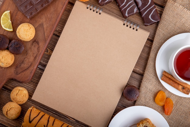 Vista superiore di vari biscotti di pasticceria dolci dello sketchbook e una tazza di tè con i bastoni di cannella su fondo di legno