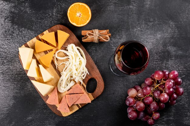 Vista superiore di vino rosso con uva, arancia e formaggio sul tagliere di legno sull'orizzontale di superficie scura