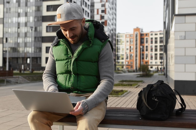 Vista urbana di elegante giovane maschio con la barba lunga che legge notizie, controlla la posta elettronica o digita un messaggio online utilizzando un computer portatile all'aperto, seduto su una panchina con borsa, viaggiando da solo, godendo del wifi gratuito