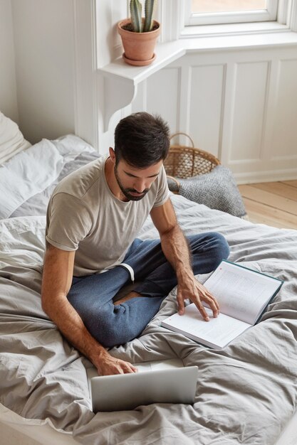 Vista verticale dall'alto di un ragazzo con la barba lunga impegnato a fare la carta del corso, studia letteratura e lavora su computer portatile