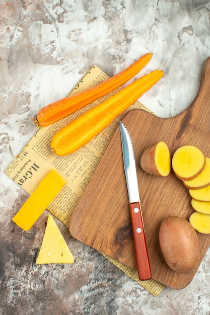 Vista verticale del fondo di cottura con varie verdure e due tipi di coltello da formaggio su tagliere di legno su un vecchio giornale su sfondo a colori misti