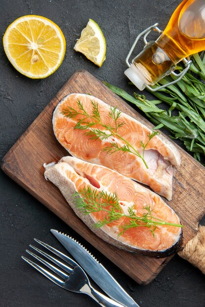 Vista verticale del pesce salmone su tagliere di legno marrone con fette di limone verde bottiglia di olio caduta sul lato destro sul tavolo scuro