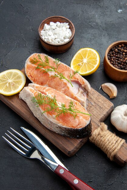 Vista verticale del pesce salmone su tagliere di legno marrone con fette di limone verde, peperoni sale, posate su tavola scura