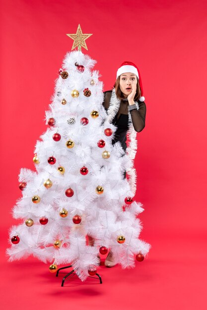 Vista verticale dell'umore del nuovo anno con una bella ragazza in un abito nero con il cappello di Babbo Natale che si nasconde dietro l'albero di Natale