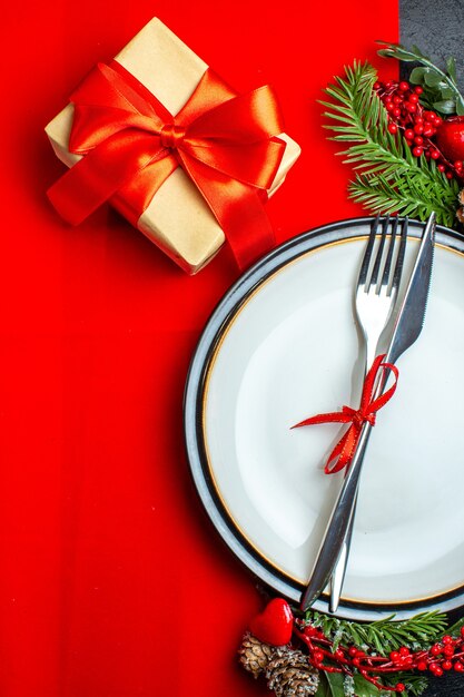 Vista verticale dello sfondo di Natale con set di posate con nastro rosso su un piatto da pranzo accessori per la decorazione rami di abete accanto a un regalo su un tovagliolo rosso