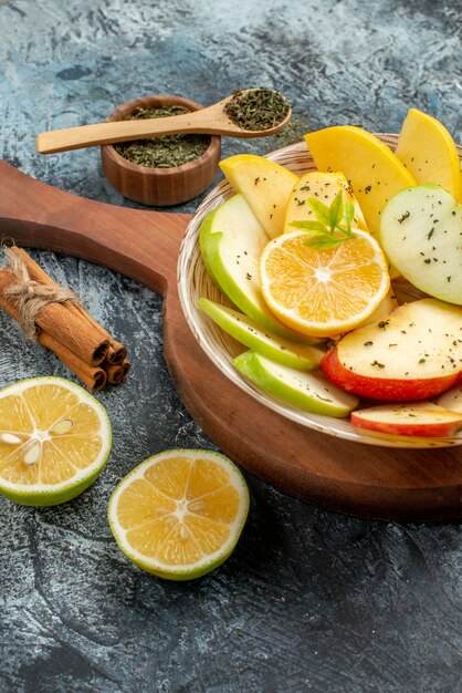 Vista verticale di fette di mela rossa verde gialla fresca su un piatto bianco con limone su un tagliere di legno lime di cannella su sfondo grigio